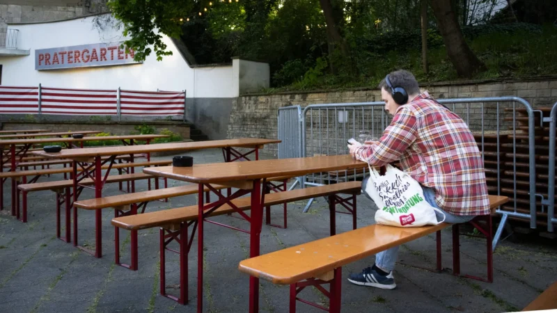 Muriel · 30/04/2026 · 2048 x 1152 Cette photographie montre une personne assise seule à une table de pique-nique en bois, absorbée par son téléphone. Le sujet principal est cette personne, vêtue d'un gilet rouge et noir à carreaux.La composition met en valeur les tables vides autour d'elle, créant une ambiance de solitude. Le cadrage est serré sur la personne, avec un arrière-plan légèrement flou incluant une clôture et un panneau "Wassergarten".La lumière semble naturelle, probablement en journée. Les couleurs dominantes sont les tons neutres (bois, métal) et les couleurs vives du gilet de la personne. Le style est réaliste et documentaire, évoquant une ambiance calme et mélancolique.