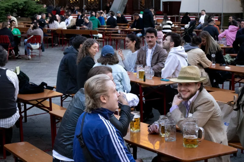 Muriel · 30/04/2026 · 2048 x 1365 Cette photographie capture un groupe de personnes en train de discuter dans un cadre extérieur convivial, probablement une brasserie ou un jardin public. Le sujet principal semble être les interactions sociales entre les convives.La composition montre plusieurs tables en bois occupées par des individus assis, certains en conversation. Le cadrage est serré sur un groupe au premier plan, tandis que l’arrière-plan reste flou mais visible.La lumière est naturelle, probablement celle d’un jour ensoleillé. Les couleurs dominantes sont des tons chauds et naturels : bois, verts, gris et quelques touches de couleurs vives comme les vêtements des personnes.L’ambiance est détendue et sociale, évoquant une journée de détente entre amis ou collègues.