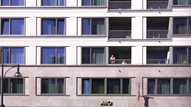 Muriel · 01/05/2026 · 2048 x 1152 Cette photographie met en scène une façade d’immeuble moderne. Le sujet principal est une personne assise sur un balcon, légèrement floutée, suggérant une vie discrète et anonyme.La composition repose sur une symétrie géométrique, avec des balcons alignés et des fenêtres rectangulaires. Le cadrage serré accentue les détails architecturaux et les jeux d’ombres.La lumière, naturelle et douce, crée des contrastes subtils entre les zones éclairées et ombragées. Les couleurs dominantes sont des tons neutres : gris, beige et blanc, avec des touches de vert (végétation).Le style est épuré et contemporain, évoquant une ambiance calme et urbaine.