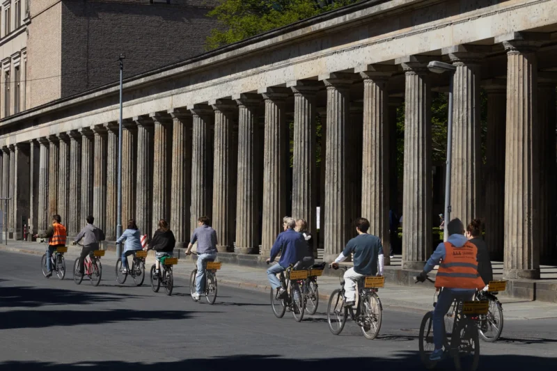 Muriel · 01/05/2026 · 2048 x 1365 Cette photographie montre un groupe de cyclistes roulant en file sur une voie urbaine. Le **sujet principal** est ce groupe, encadré par une **composition** horizontale mettant en valeur une colonnade classique en arrière-plan.Le **cadrage** est serré sur les cyclistes, avec une perspective légèrement basse. La **lumière** est naturelle et vive, créant des contrastes nets. Les **couleurs dominantes** sont neutres (gris des bâtiments, noir des vélos) avec des touches vives (vêtements colorés des cyclistes).Le **style** est documentaire et dynamique, évoquant une **ambiance** urbaine moderne et active.