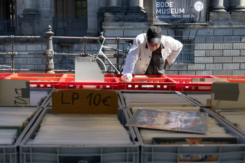 Muriel · 01/05/2026 · 2048 x 1365 Cette photographie montre un homme en train de manipuler une longue règle métallique rouge marquée "LP 10€". Le sujet principal est cet homme, vêtu d’une combinaison blanche, dans un cadre industriel ou artistique.La composition met en avant un espace structuré avec des éléments métalliques et des boîtes. Le cadrage est serré, centré sur l’action. La lumière semble naturelle, bien répartie, éclairant distinctement la scène. Les couleurs dominantes sont les tons froids (gris, blanc) et le rouge vif de la règle.L’ambiance est à la fois technique et artistique, évoquant un atelier ou une installation. Le style est documentaire et réaliste.