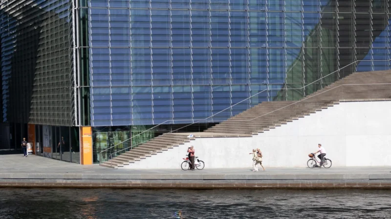 Muriel · 01/05/2026 · 2048 x 1152 Cette photographie met en scène trois cyclistes comme sujets principaux, évoluant sur une promenade en bordure d’eau. La composition joue sur les lignes droites des bâtiments modernes et des escaliers en béton, créant un contraste géométrique. Le cadrage serré isole les personnages et la structure architecturale, avec une perspective légèrement basse.La lumière est naturelle et douce, reflétée par l’eau. Les couleurs dominantes sont les tons froids (bleu du bâtiment, gris du béton) et chauds (teintes des vêtements des cyclistes). Le style est épuré et contemporain, évoquant une ambiance urbaine paisible et moderne.