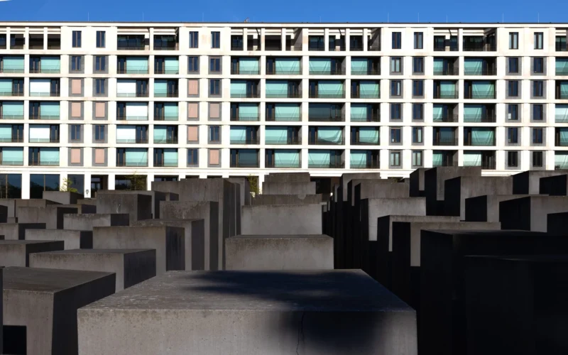 Muriel · 01/05/2026 · 2048 x 1280 Cette photographie met en scène un mémorial composé de blocs de béton disposés en grille régulière, appelé *Memorial to the Murdered Jews of Europe* (Berlin). Le sujet principal est cette installation minimaliste et solennelle.La composition joue sur les lignes droites et la répétition géométrique, renforcée par le cadrage serré. La lumière, naturelle et contrastée, accentue les ombres et les formes anguleuses. Les couleurs dominantes sont les tons gris-beige des blocs et le blanc des bâtiments en arrière-plan.Le style est épuré et symbolique, créant une ambiance à la fois austère et poétique, évoquant la mémoire et la réflexion.