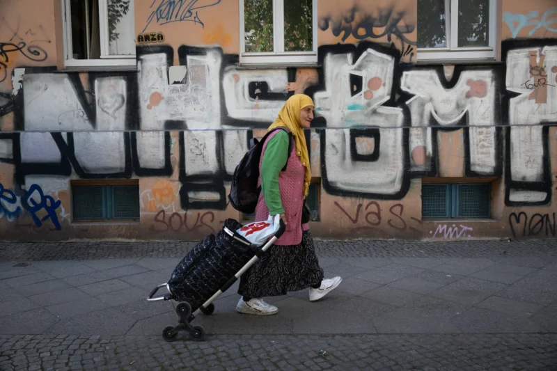 Muriel · 02/05/2026 · 2048 x 1365 Cette photographie met en scène une femme vêtue d’un hijab jaune et d’une veste colorée, poussant un landau. Le sujet principal est centré, avec une composition simple et équilibrée.Le cadrage est serré, mettant l’accent sur la personne et son environnement immédiat. La lumière est naturelle, légèrement tamisée, créant des ombres douces. Les couleurs dominantes sont les tons vifs de ses vêtements (jaune, vert, rouge) contrastant avec les teintes grises et les graffitis colorés du mur en arrière-plan.Le style est documentaire et réaliste, évoquant une ambiance urbaine et quotidienne, presque intimiste malgré le contexte public.