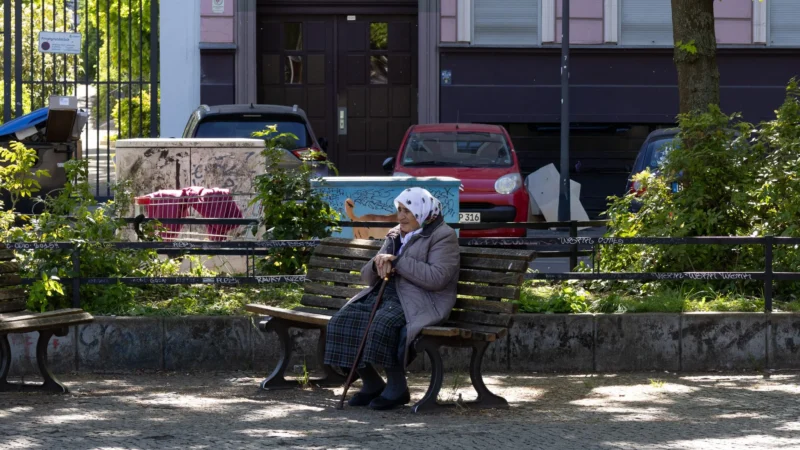 Muriel · 02/05/2026 · 2048 x 1152 Cette photographie met en scène une personne âgée assise sur un banc, vêtue d’un chapeau et d’un manteau, semblant observer son environnement. La composition est simple et naturelle, avec un cadrage serré sur le sujet et un arrière-plan légèrement flou.Les couleurs dominantes sont des tons sobres : gris, vert des plantes, et touches de rouge et bleu des véhicules. La lumière semble douce et naturelle, probablement en journée. Le style est réaliste et documentaire, capturant un moment de calme. L’ambiance est paisible et introspective, évoquant la sérénité du quotidien urbain.