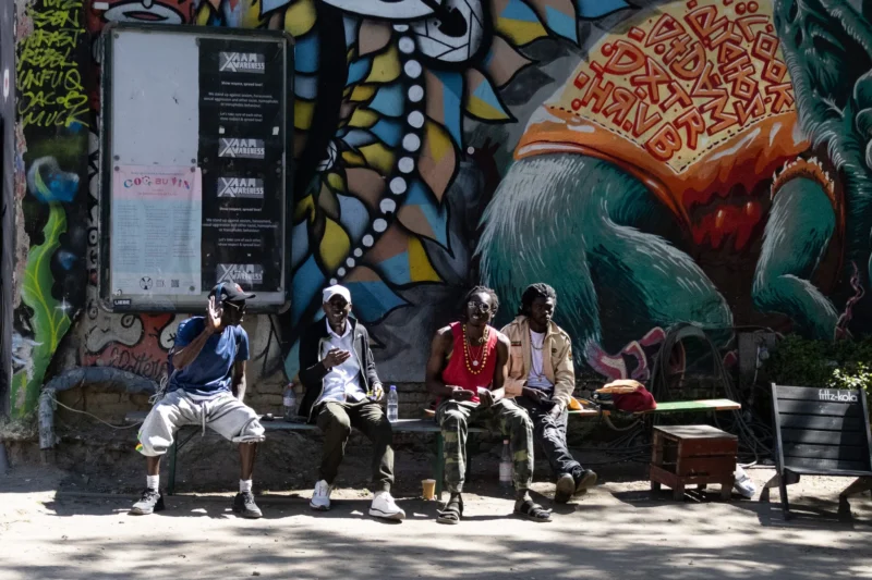 Muriel · 02/05/2026 · 2048 x 1365 Cette photographie capture un groupe de personnes assises sur un banc public devant un mur coloré. Le **sujet principal** est ce groupe, dont les visages sont floutés, créant une ambiance anonyme et universelle.La **composition** est équilibrée, avec le banc et les personnages centrés. Le **cadrage** est serré, mettant en valeur les détails du mur et les interactions discrètes des individus. La **lumière** semble naturelle, probablement en journée, avec des ombres douces.Les **couleurs dominantes** sont vives et contrastées : tons chauds (oranges, jaunes) et froids (bleus, verts) sur le mur mural, tandis que les vêtements des personnes sont neutres. Le **style** est documentaire et réaliste, évoquant une scène de vie quotidienne. L’**ambiance** est calme et contemplative.
