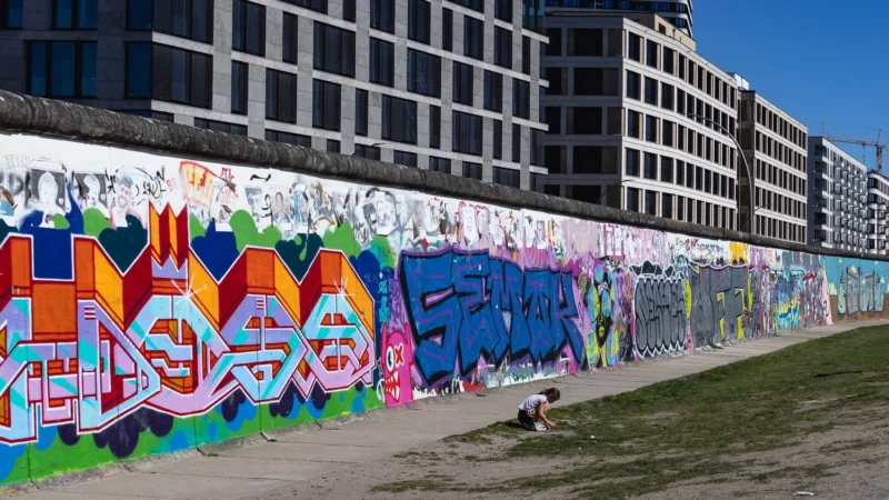Muriel · 02/05/2026 · 2048 x 1152 Cette photographie montre un mur couvert de **graffitis colorés**, sujet principal de l'image. La composition juxtapose ces fresques urbaines avec des bâtiments modernes en arrière-plan. Le cadrage est horizontal, capturant une longue portion du mur.La lumière est naturelle, probablement ensoleillée, mettant en valeur les couleurs vives. Les **couleurs dominantes** sont le bleu, le rose, le vert et le jaune, créant un contraste dynamique. Le style est **urbain et artistique**, reflétant la culture street art. L'ambiance est énergique et créative, mêlant art et architecture contemporaine.
