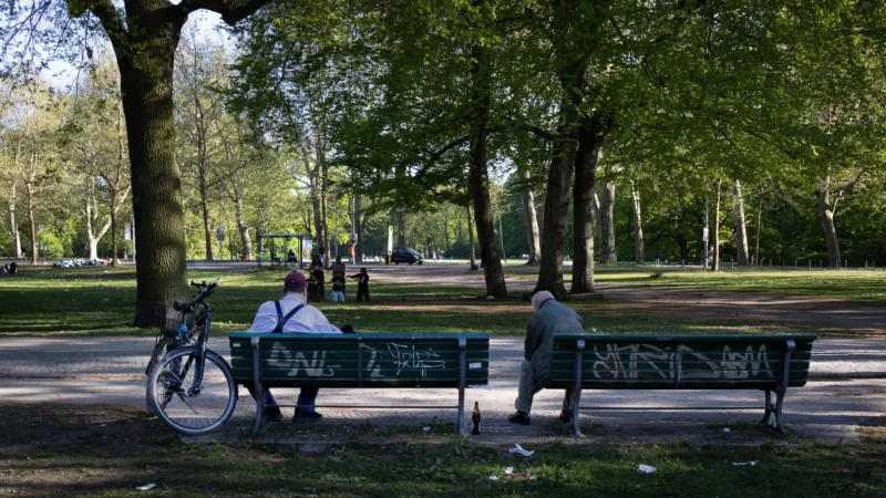 Muriel · 02/05/2026 · 2048 x 1152 Cette photographie capture un parc verdoyant où le sujet principal est un banc en métal avec une inscription. Deux personnes, dont une avec un vélo, sont assises. La composition est équilibrée, avec un cadrage large incluant des arbres et une pelouse.La lumière est naturelle et douce, typique d’un jour ensoleillé. Les couleurs dominantes sont les verts des arbres et de l’herbe, avec des touches de gris métallique du banc. Le style est réaliste et documentaire, reflétant une scène de vie quotidienne. L’ambiance est paisible et sereine, évoquant la détente et la tranquillité.