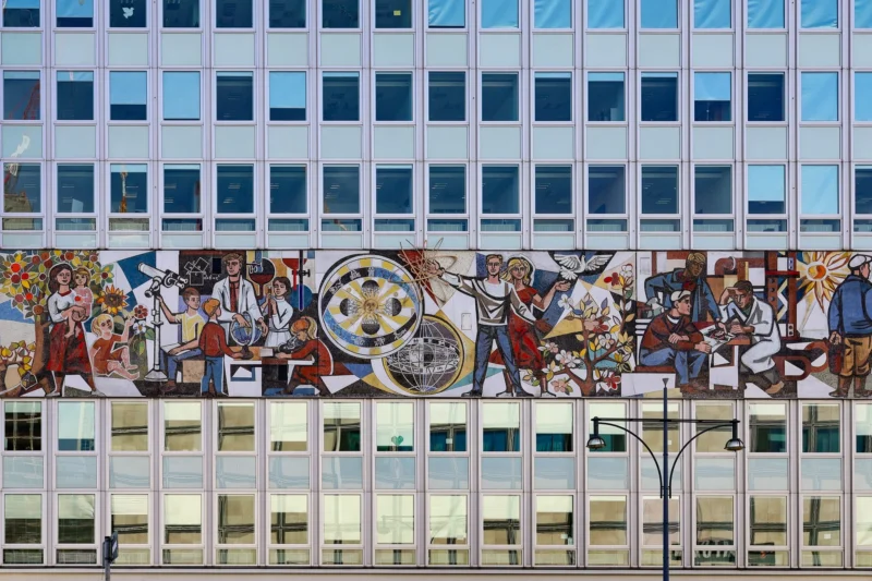 Muriel · 03/05/2026 · 2048 x 1365 Cette photographie montre un **mural coloré** sur la façade d’un immeuble moderne. Le **sujet principal** est une fresque représentant des scènes historiques et culturelles, avec des personnages variés et un cadran central évoquant une horloge.La **composition** est horizontale, bien cadrée sur le bâtiment. La **lumière** est naturelle, éclairant uniformément la scène. Les **couleurs dominantes** sont vives : ocres, rouges, bleus et verts.Le **style** est réaliste et détaillé, mêlant art mural et architecture contemporaine. L’**ambiance** est à la fois dynamique et historique, reflétant une célébration culturelle en milieu urbain.