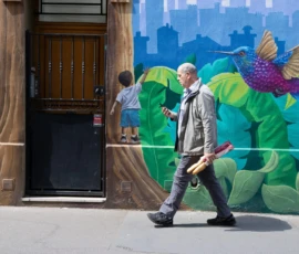 Muriel · 09/05/2026 · 2048 x 1365 Cette photographie met en scène un homme âgé marchant devant un mur coloré. Le sujet principal est cet homme, vêtu simplement, en mouvement.La composition joue sur le contraste entre le réalisme de l’homme et l’abstraction végétale du mural. Le cadrage est serré, centré sur le sujet et le mur, avec une profondeur limitée.La lumière est naturelle et claire, créant des ombres douces. Les couleurs dominantes sont les verts, bleus et touches de violet du mural, apportant une ambiance vibrante et joyeuse. Le style est urbain et artistique, mêlant vie quotidienne et expression créative.