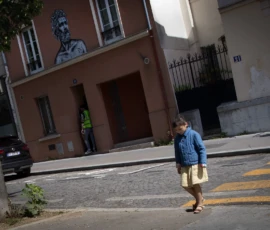 Muriel · 09/05/2026 · 2048 x 1364 Cette photographie met en scène un enfant comme sujet principal, marchant sur un trottoir. La composition est simple, avec un cadrage serré sur l’enfant, isolé au premier plan. En arrière-plan, un mur avec un graffiti représentant un visage et une personne en vert ajoutent du contraste.La lumière semble naturelle, légèrement tamisée, créant des ombres douces. Les couleurs dominantes sont les tons neutres (gris du trottoir, beige des bâtiments) et quelques touches vives (bleu du vêtement de l’enfant, vert de la personne en arrière-plan).L’ambiance est calme et un peu mélancolique, renforcée par les tons sobres et le jeu d’ombres.