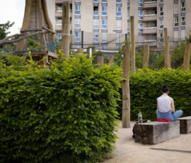 Muriel · 09/05/2026 · 2048 x 1365 Cette photographie montre une personne assise sur un banc en bois, entourée de végétation dense. Le sujet principal est cette personne, vêtue de blanc, en position méditative ou contemplative.La composition est simple et épurée, avec un cadrage serré sur le premier plan. En arrière-plan, des bâtiments modernes et des structures architecturales sont visibles, créant un contraste entre nature et urbanité. La lumière semble naturelle, douce, et légèrement tamisée, suggérant un moment calme, probablement en journée.Les couleurs dominantes sont les verts profonds des plantes, les tons neutres du bois et du béton, et les blancs des vêtements de la personne. L’ambiance est paisible et introspective.