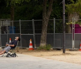 Muriel · 09/05/2026 · 2048 x 1152 Cette photographie montre un individu assis sur un trottinette électrique, sujet principal en position centrale. La composition met en avant un espace urbain avec une clôture métallique et des cônes orange. Le cadrage est serré, centré sur le sujet et son environnement immédiat.La lumière semble naturelle, avec des ombres marquées, suggérant un jour ensoleillé. Les couleurs dominantes sont les tons neutres (gris de la clôture, beige du sol) et les touches vives des cônes orange. Le style est documentaire et réaliste, capturant une scène du quotidien. L’ambiance est calme et légèrement mélancolique.