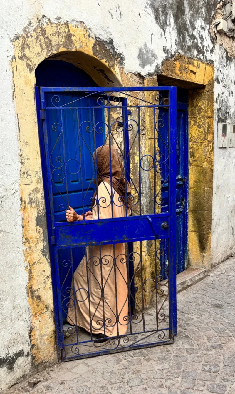 Sophie · 2692 x 4507 Cette photographie montre une personne vêtue d’une robe traditionnelle, partiellement cachée derrière une porte ou une fenêtre à grille bleue. Le sujet principal est flou, suggérant un mouvement ou une intention de discrétion.La composition est centrée sur cette ouverture, encadrée par des murs blancs et une architecture rustique. Le cadrage est serré, mettant en valeur la grille et le personnage.Les couleurs dominantes sont le bleu vif de la grille, le beige de la robe et le blanc des murs. La lumière semble naturelle, créant des contrastes doux. L’ambiance est à la fois mystérieuse et chaleureuse, évoquant un lieu traditionnel et authentique.