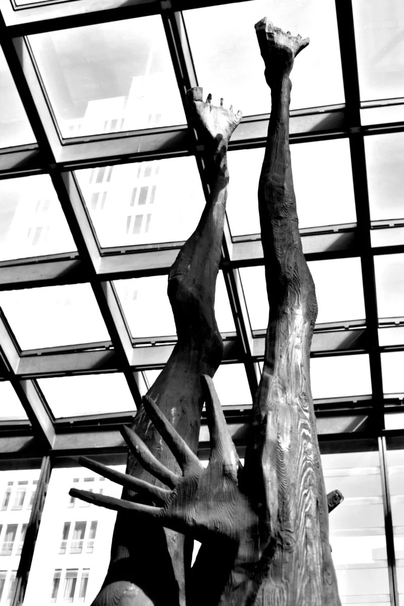 Fabienne · 30/04/2026 · 3662 x 5488 Cette photographie en noir et blanc met en scène une sculpture abstraite représentant une main et un bras émergent d’un tronc d’arbre stylisé. La composition joue sur les contrastes entre les formes organiques et les lignes géométriques de la structure vitrée en arrière-plan. Le cadrage serré isole le sujet, accentuant son expression dramatique. La lumière, naturelle et filtrée, crée des ombres marquées, renforçant le côté sculptural et presque spectral de l’œuvre. Les tons dominants, du noir au gris, confèrent une ambiance mystérieuse et contemplative, typique du style artistique moderne et expressionniste.