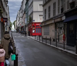 Muriel · 16/05/2026 · 2048 x 1365 Cette photographie montre une rue urbaine déserte avec une femme en manteau marron et chapeau comme sujet principal, marchant avec un sac vert. La composition met en valeur une perspective linéaire, avec des bâtiments alignés de chaque côté.Le cadrage est serré, guidant le regard vers le fond de la rue. La lumière est naturelle mais tamisée, créant une ambiance sobre et légèrement mélancolique. Les couleurs dominantes sont les tons gris-beige des bâtiments, rehaussés par le rouge vif du téléphone public et les touches de vert et marron des vêtements.Le style est réaliste et documentaire, évoquant une atmosphère calme et presque intemporelle.