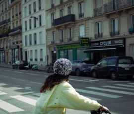 Muriel · 16/05/2026 · 2048 x 1463 Cette photographie montre une femme, vue de dos, traversant un carrefour à vélo. Elle porte un manteau jaune pâle et un chapeau orné.La composition est dynamique, avec une perspective légèrement basse. Le cadrage met en valeur la scène urbaine, incluant des bâtiments, des voitures et des enseignes.La lumière est naturelle, probablement en journée, avec des tons doux. Les couleurs dominantes sont les jaunes, gris et verts, créant une ambiance urbaine paisible et légèrement rétro. Le style semble documentaire, capturant un moment de vie quotidienne.