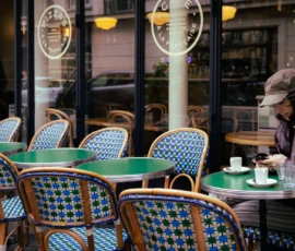 Muriel · 16/05/2026 · 2048 x 1365 Cette photographie montre une terrasse de café parisienne. Le sujet principal est une femme assise à une table, vêtue d’un manteau et d’un chapeau, en train de boire. La composition met en valeur des tables et chaises typiques en métal et carreaux bleus et blancs. Le cadrage est serré, centré sur la scène intérieure et extérieure.La lumière est douce et tamisée, créant une ambiance chaleureuse et intimiste. Les couleurs dominantes sont les tons neutres (beige, gris) et les bleus pastel des chaises. Le style évoque un cadre classique et élégant, typique des cafés parisiens. L’ambiance est paisible et romantique.