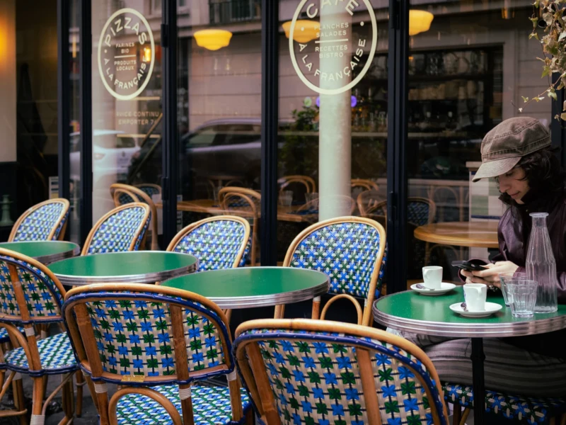 Muriel · 16/05/2026 · 2048 x 1365 Cette photographie montre une terrasse de café parisienne. Le sujet principal est une femme assise à une table, vêtue d’un manteau et d’un chapeau, en train de boire. La composition met en valeur des tables et chaises typiques en métal et carreaux bleus et blancs. Le cadrage est serré, centré sur la scène intérieure et extérieure.La lumière est douce et tamisée, créant une ambiance chaleureuse et intimiste. Les couleurs dominantes sont les tons neutres (beige, gris) et les bleus pastel des chaises. Le style évoque un cadre classique et élégant, typique des cafés parisiens. L’ambiance est paisible et romantique.