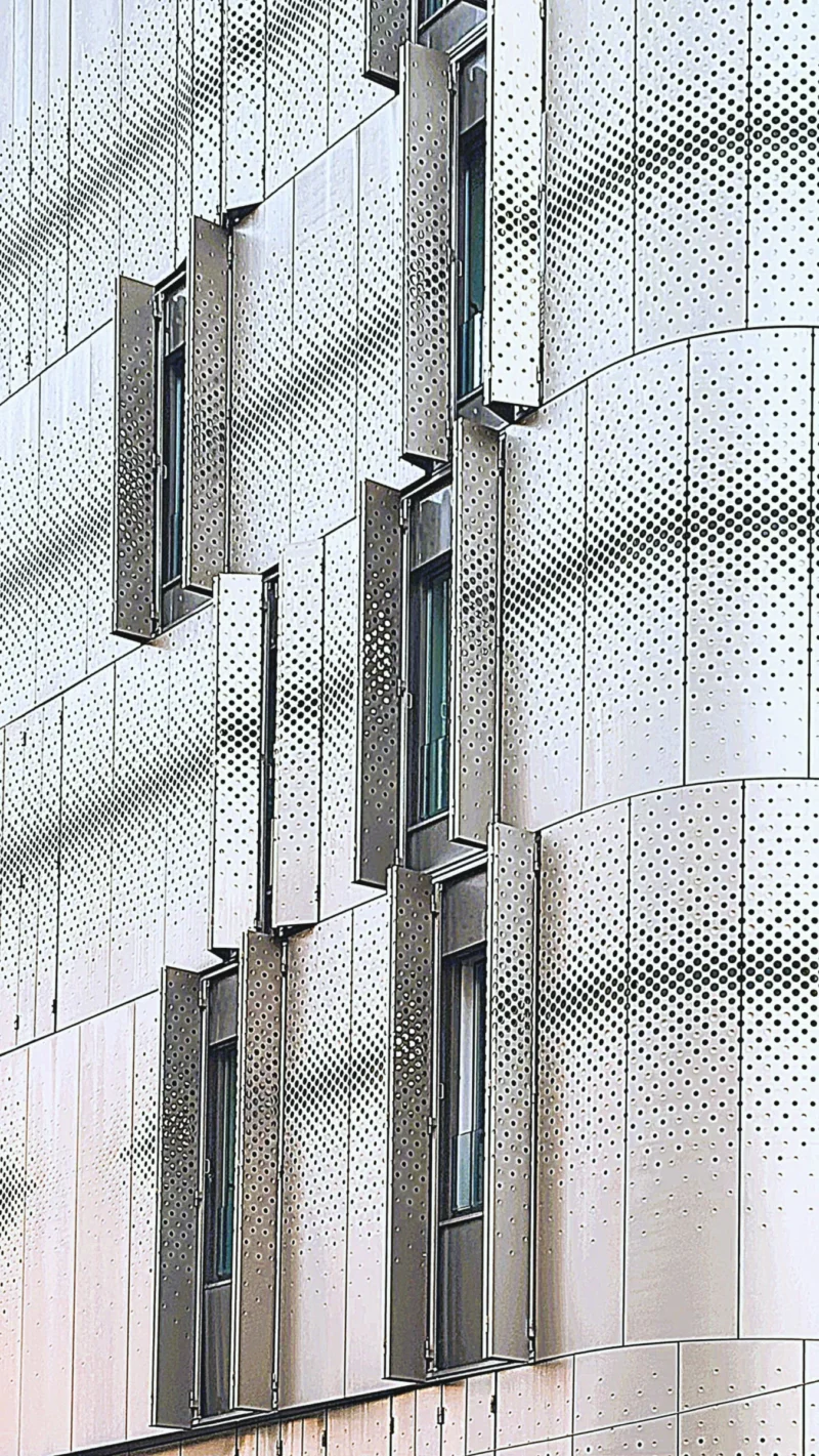 Sophie · 1736 x 3085 Cette photographie met en scène une façade architecturale moderne. Le sujet principal est une structure en béton et métal avec des fenêtres verticales étroites et des éléments en treillis métallique.La composition repose sur des lignes verticales et horizontales, créant un jeu géométrique. Le cadrage serré accentue les détails architecturaux. La lumière, naturelle et douce, éclaire uniformément la surface, mettant en valeur les textures.Les couleurs dominantes sont des tons neutres : gris clair, beige et blanc. Le style est épuré et contemporain, évoquant une ambiance minimaliste et industrielle.