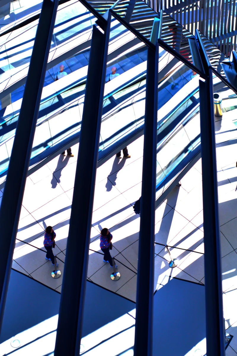 Fabienne · 01/05/2026 · 3662 x 5488 Cette photographie capture un espace urbain moderne vu d’en haut. Le sujet principal est une passerelle ou un pont piétonnier avec des silhouettes de personnes en mouvement. La composition repose sur des lignes verticales et horizontales, créées par les structures métalliques et les bandes bleues du sol.Le cadrage est serré, mettant en valeur les jeux d’ombres et de répétitions. La lumière est naturelle et vive, accentuant les contrastes. Les couleurs dominantes sont le bleu, le gris et le noir, donnant un aspect contemporain et épuré. L’ambiance est dynamique et architecturale, reflétant une ville moderne et fonctionnelle.