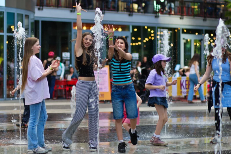 Fabienne · 01/05/2026 · 3816 x 2544 Cette photographie capture un groupe de jeunes femmes en pleine fête près d'une fontaine à jets d’eau. Le sujet principal est une jeune femme au centre, levant les bras joyeusement.La composition est dynamique, avec un cadrage serré sur les personnages. La lumière est naturelle, probablement en soirée, avec des éclairages urbains qui ajoutent une ambiance festive. Les couleurs dominantes sont vives : tons pastel (violet, bleu) et contrastes avec les vêtements colorés.Le style est spontané et joyeux, évoquant une ambiance détendue et conviviale, typique d’une soirée estivale en ville.