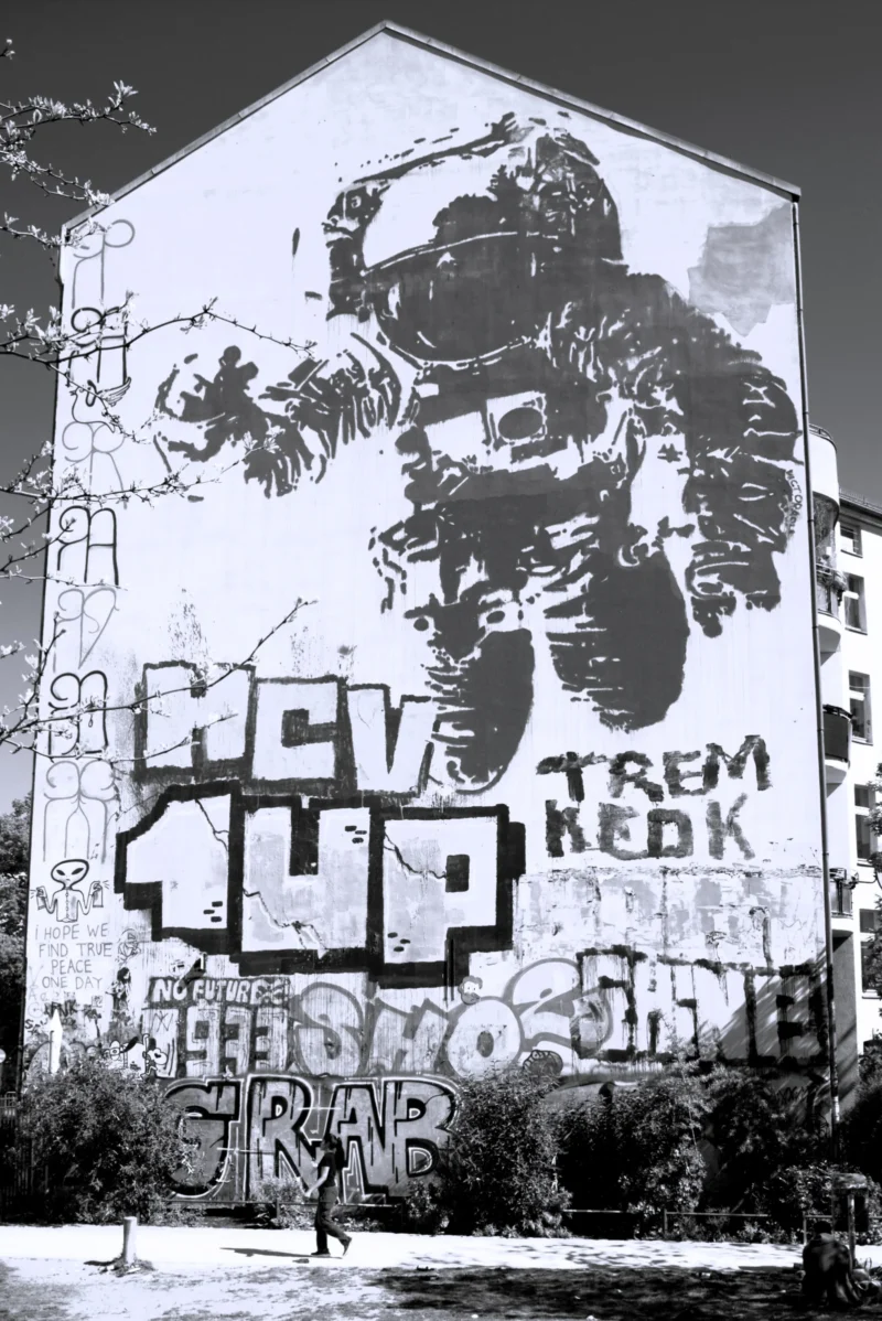 Fabienne · 2838 x 4250 Cette photographie montre un graffiti mural en noir et blanc. Le sujet principal est une illustration stylisée d’un personnage en position dynamique, entourée de tags et de textes comme "MC UP", "TREM NEOK" et "SORAB". La composition est verticale, avec le graffiti couvrant presque toute la hauteur du bâtiment.Le cadrage est serré, mettant en valeur les détails du mur. La lumière semble naturelle, sans ombres marquées, ce qui accentue les contrastes des traits. Les couleurs dominantes sont les nuances de gris, typiques d’une photo en noir et blanc. L’ambiance est urbaine et artistique, reflétant la culture street art.