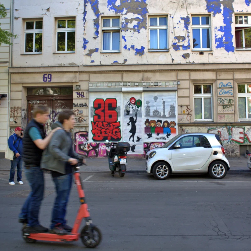 Fabienne · 02/05/2026 · 3385 x 3385 Cette photographie capture une scène urbaine dynamique. Le sujet principal est un homme sur un trottinette électrique rouge, au premier plan. La composition montre une rue animée avec des passants, des graffitis colorés sur un mur dégradé et un véhicule compact blanc.Le cadrage est serré sur le trottinettiste, avec une profondeur de champ qui inclut les éléments en arrière-plan. La lumière semble naturelle, probablement en journée, avec des ombres douces. Les couleurs dominantes sont les tons froids (bleu, gris) contrastant avec les touches chaudes (rouge de la trottinette, rouge des graffitis).L’ambiance est moderne et vivante, reflétant un quartier urbain en mouvement.