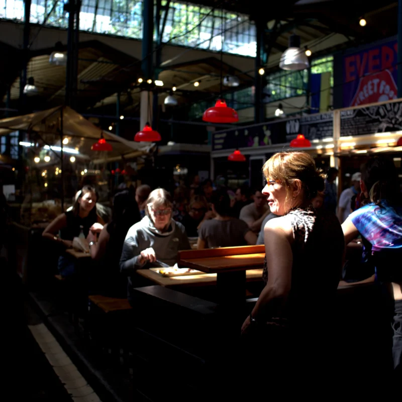 Fabienne · 02/05/2026 · 3428 x 3428 Cette photographie capture une scène animée dans un bar ou un marché couvert. Le sujet principal est une femme assise à une table, tournée vers la gauche, observant les gens autour d'elle.La composition montre plusieurs personnes en interaction, créant une ambiance conviviale. Le cadrage est serré, mettant en valeur les détails des visages et des objets.La lumière est tamisée, avec des éclairages chauds provenant de suspensions rouges. Les couleurs dominantes sont les tons sombres et chauds, avec des éclats de rouge et de bleu.Le style est réaliste et spontané, évoquant une ambiance nocturne et vivante.
