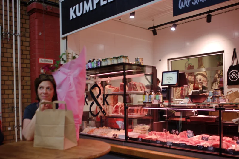 Fabienne · 02/05/2026 · 4207 x 2805 Cette photographie montre un comptoir de charcuterie dans une boutique nommée "Kumpele". Le sujet principal est une vitrine éclairée mettant en avant des produits de viande et fromage. À gauche, une affiche promotionnelle représente une femme tenant un sac avec un ballon rose.La composition est symétrique, avec un cadrage serré sur le comptoir. La lumière est artificielle et met en valeur les produits. Les couleurs dominantes sont les tons chauds (rouges, bruns) et les touches froides (bleu du fond). L’ambiance est chaleureuse et professionnelle, typique d’un commerce alimentaire haut de gamme.