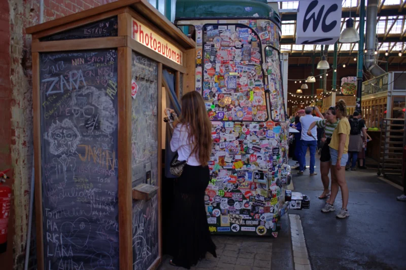 Fabienne · 02/05/2026 · 5203 x 3469 Cette photographie montre une jeune femme utilisant une cabine téléphonique vintage couverte de graffitis et de stickers. Le sujet principal est cette cabine, entourée d’éléments urbains colorés.La composition est dynamique, avec un cadrage serré sur la cabine et des personnes en arrière-plan. La lumière est naturelle, probablement en journée, éclairant les tons chauds et froids. Les couleurs dominantes sont vives et variées, avec des touches de rouge, bleu et jaune.Le style est urbain et artistique, évoquant une ambiance créative et nostalgique, typique d’un quartier animé et coloré.