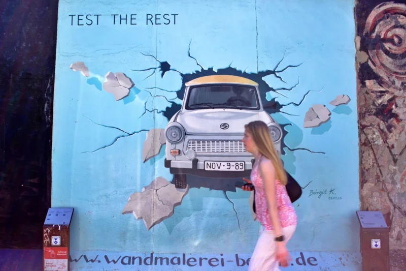 Fabienne · 02/05/2026 · 4910 x 3274 Cette photographie montre une femme posant devant une fresque murale. Le sujet principal est un camion Volkswagen intégré dans une composition artistique colorée et fragmentée, évoquant un style street art ou graffiti.Le cadrage est serré sur la fresque et la femme, avec des éléments textuels ("TEST THE REST", "Wandmalerei-br..."). Les couleurs dominantes sont le bleu, le blanc et des touches de rose et jaune. La lumière semble naturelle, mettant en valeur les détails du mur.L’ambiance est dynamique et créative, mêlant art urbain et interaction humaine.