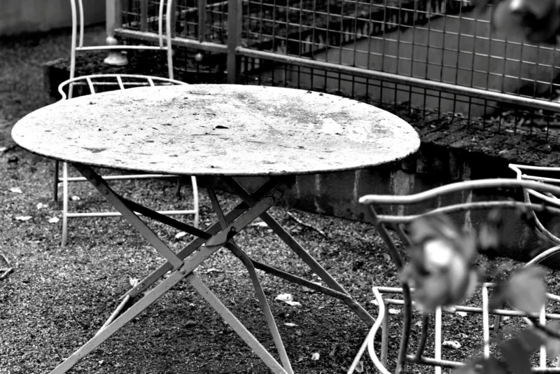 Fabienne · 17/05/2026 · 5488 x 3662 Cette photographie en noir et blanc met en scène un **tabouret et une table de café** métalliques, légèrement usés, posés sur un sol pavé.Le **sujet principal** est la table ronde, entourée de chaises, avec une légère accumulation de poussière. La **composition** est simple et épurée, avec un **cadrage serré** qui isole ces objets.La **lumière** semble naturelle, douce, créant des contrastes subtils. Les **couleurs dominantes** sont les nuances de gris, renforçant une ambiance **nostalgique et mélancolique**. Le style est **minimaliste et documentaire**, évoquant une scène abandonnée ou un lieu peu fréquenté.