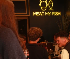 Sophie · 3545 x 2848 Cette photographie montre un groupe de personnes dans un restaurant nommé **« Meat My Fish »**, visible grâce à une enseigne néon. Le sujet principal semble être l’ambiance conviviale et détendue autour d’un repas.La composition met en avant les visages des convives, légèrement floutés, avec un cadrage serré sur eux. La lumière est tamisée, créant une ambiance chaleureuse et intimiste. Les couleurs dominantes sont les tons chauds (oranges, jaunes) et les teintes douces des vêtements.Le style est informel et moderne, évoquant une ambiance décontractée et amicale dans un cadre urbain ou branché.