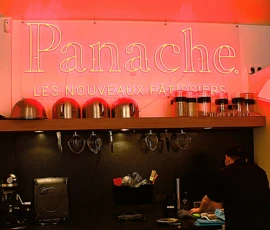 Sophie · 3026 x 2267 Cette photographie montre l'intérieur d'un café ou restaurant nommé **Panache**, avec pour slogan *"Les Nouveaux Artisans"*.Le **sujet principal** est l'arrière du comptoir, où l'on voit des ustensiles de cuisine et des objets divers. La **composition** est symétrique avec des éléments alignés sur une étagère. Le **cadrage** est serré, mettant en valeur les détails du comptoir.La **lumière** est tamisée, avec des éclairages chauds et une ambiance rougeâtre. Les **couleurs dominantes** sont le rouge, le noir et le blanc. Le style est moderne et épuré, créant une **ambiance** chaleureuse et artisanale.