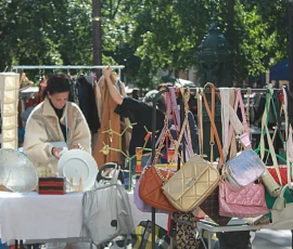 Sophie · 4272 x 2848 Cette photographie montre un marché en plein air. Le sujet principal est une vendeuse derrière un étalage de divers accessoires : sacs, bijoux et objets décoratifs.La composition est dynamique, avec des tables chargées de marchandises disposées en désordre organisé. Le cadrage est serré sur l’étal, avec des éléments en arrière-plan floutés, comme des arbres et d’autres stands.La lumière est naturelle et douce, probablement en journée. Les couleurs dominantes sont neutres (beige, blanc) avec des touches vives (rose, vert). L’ambiance est chaleureuse et artisanale, évoquant un marché traditionnel et convivial.