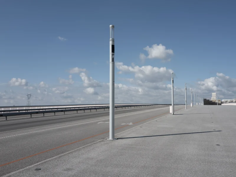 Jean-Philippe · 12/05/2026 · 7795 x 5846 Cette photographie montre un **pont autoroutier** avec des **poteaux de signalisation** comme sujet principal. La composition est épurée, avec une perspective axiale menant vers l’horizon. Le cadrage est large, capturant une portion de la route et du ciel.La lumière est naturelle, avec un éclairage doux et des ombres légères, suggérant un jour ensoleillé. Les **couleurs dominantes** sont les tons gris (béton, route) et bleu (ciel), rehaussés par des touches de blanc (nuages).Le style est **minimaliste et documentaire**, évoquant une ambiance calme et moderne, presque industrielle.