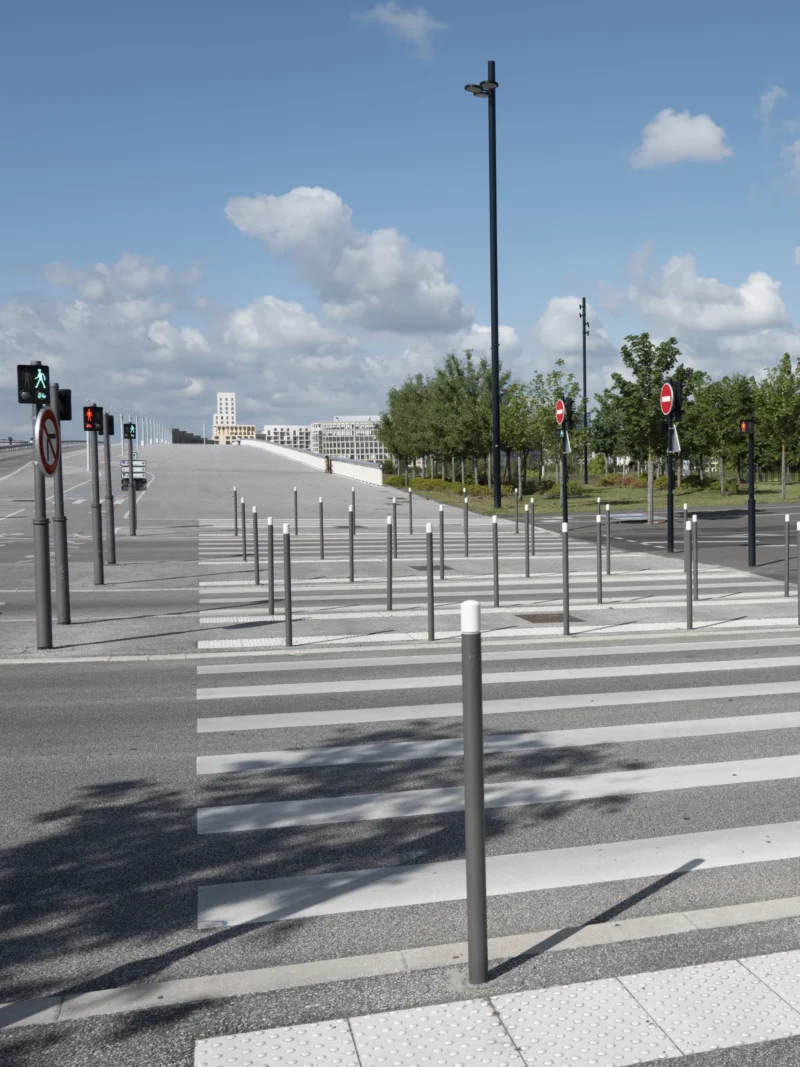 Jean-Philippe · 12/05/2026 · 4714 x 6285 Cette photographie montre un **passage piéton** urbain presque désert. Le **sujet principal** est la traversée marquée par des bandes blanches et délimitée par des **poteaux métalliques**. La composition est symétrique, avec des lignes directrices menant vers l’horizon.Le **cadrage** est serré, capturant une portion de trottoir et de route. La **lumière** est naturelle, éclatante, avec des ombres nettes, suggérant un jour ensoleillé. Les **couleurs dominantes** sont les tons gris (asphalte, poteaux) et blancs (marquages), rehaussés par le bleu du ciel et le vert des arbres en arrière-plan.Le style est **minimaliste et documentaire**, évoquant une **ambiance calme et ordonnée**.