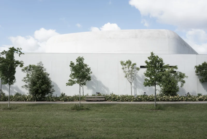 Jean-Philippe · 12/05/2026 · 7758 x 5200 Cette photographie met en scène un bâtiment moderne au design épuré et aux formes arrondies, recouvert d’un enduit blanc. Le sujet principal est ce mur lisse et imposant, presque abstrait.La composition repose sur un cadrage horizontal, avec un premier plan végétalisé (pelouse et arbres) qui crée une opposition entre nature et architecture. La lumière est naturelle, douce et légèrement diffuse, suggérant un jour nuageux. Les couleurs dominantes sont le blanc du bâtiment, le vert des arbres et le vert-gris de la pelouse, renforçant une ambiance calme et minimaliste. Le style est contemporain, épuré et architectural.