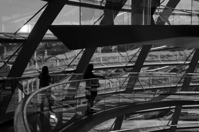Thomas · 01/05/2026 · 3080 x 2048 Cette photographie met en scène une structure architecturale moderne, probablement un hall ou un centre commercial avec des escaliers mécaniques et des passerelles métalliques. Le sujet principal est l'intersection des lignes géométriques et des jeux d'ombres.La composition repose sur des lignes diagonales et des formes répétitives, créant un effet dynamique. Le cadrage serré accentue la profondeur et la perspective. La lumière, naturelle et tamisée, génère des contrastes nets entre ombres et lumières. Les couleurs dominantes sont les tons gris et noirs, avec des touches de blanc.Le style est épuré et contemporain, évoquant une ambiance à la fois futuriste et minimaliste.