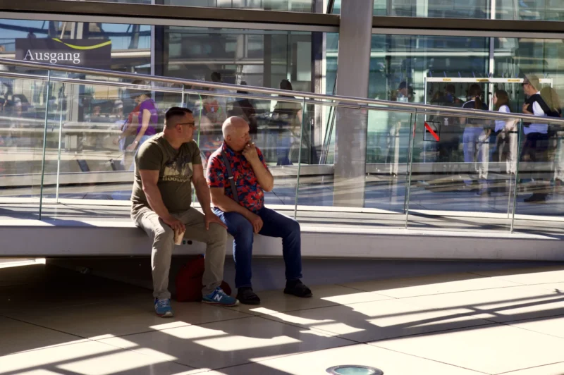 Thomas · 01/05/2026 · 3080 x 2048 Cette photographie montre deux personnes assises sur un banc dans un espace moderne, probablement une gare ou un aéroport. Le sujet principal est ces deux individus, l’un avec un sac à roulettes.La composition est simple et naturelle, avec un cadrage serré sur les deux personnes. L’arrière-plan, flou et lumineux, met en valeur les sujets. La lumière est abondante et naturelle, créant des contrastes doux.Les couleurs dominantes sont neutres (gris, beige) avec des touches de rouge et bleu sur les vêtements. Le style est réaliste et documentaire, capturant un moment du quotidien. L’ambiance est calme et détendue.