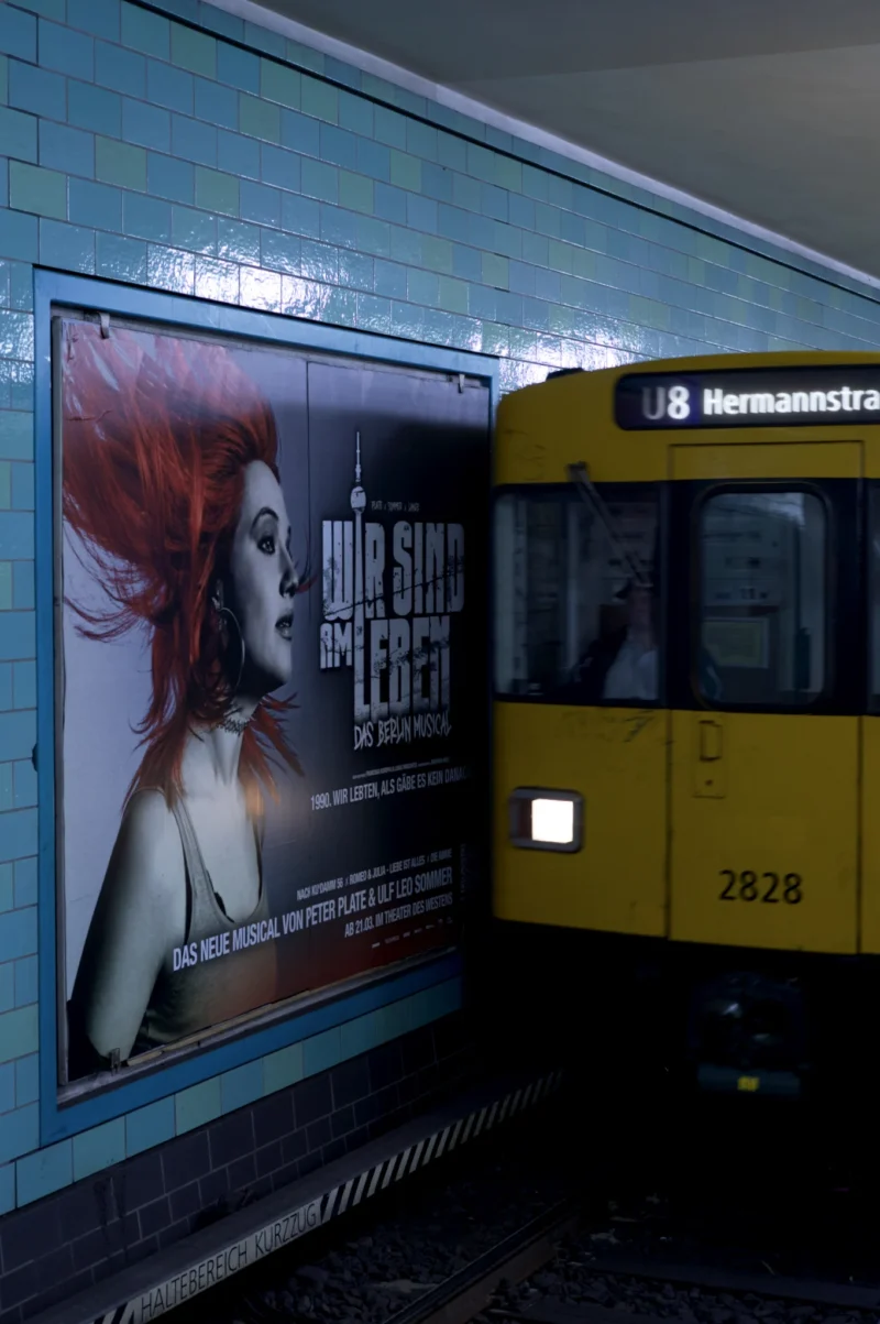 Thomas · 02/05/2026 · 1362 x 2048 Cette photographie montre un tramway jaune à l'arrêt, avec une affiche publicitaire pour une marque de parfum "Shiseido" en arrière-plan. Le sujet principal est l'affiche représentant une femme aux cheveux rouges.La composition est simple, avec un cadrage serré sur le côté du tramway et l'affiche. La lumière est tamisée, créant une ambiance nocturne ou intérieure peu éclairée. Les couleurs dominantes sont le bleu-gris de la station, le jaune vif du tramway, et les tons rouges et blancs de l'affiche. Le style est urbain et moderne, évoquant une ambiance contemporaine et dynamique.