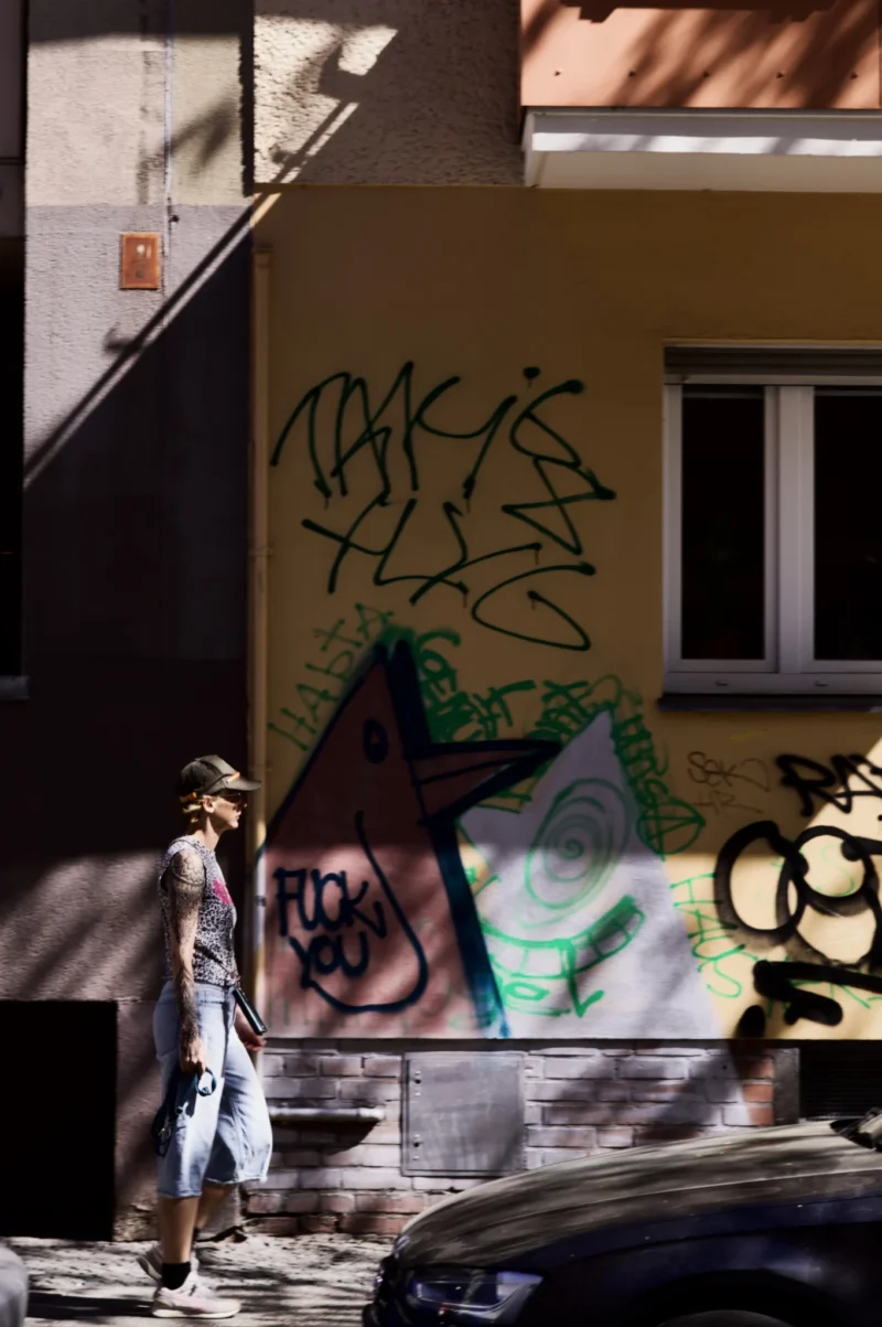 Thomas · 02/05/2026 · 1362 x 2048 Cette photographie capture une scène urbaine avec une personne en mouvement comme sujet principal, vêtue d'une veste claire et d'un chapeau. La composition met en valeur un graffiti stylisé sur un mur jaune, avec des éléments architecturaux comme une rampe d’escalier et des voitures en arrière-plan.Le cadrage est dynamique, légèrement flou, suggérant un mouvement rapide. La lumière naturelle, vive et contrastée, éclaire la scène, créant des ombres marquées. Les couleurs dominantes sont les tons jaunes, verts et gris, avec des touches de bleu et de noir dans les graffitis.L’ambiance est urbaine et vivante, mêlant art de rue et quotidien. Le style est réaliste et spontané.