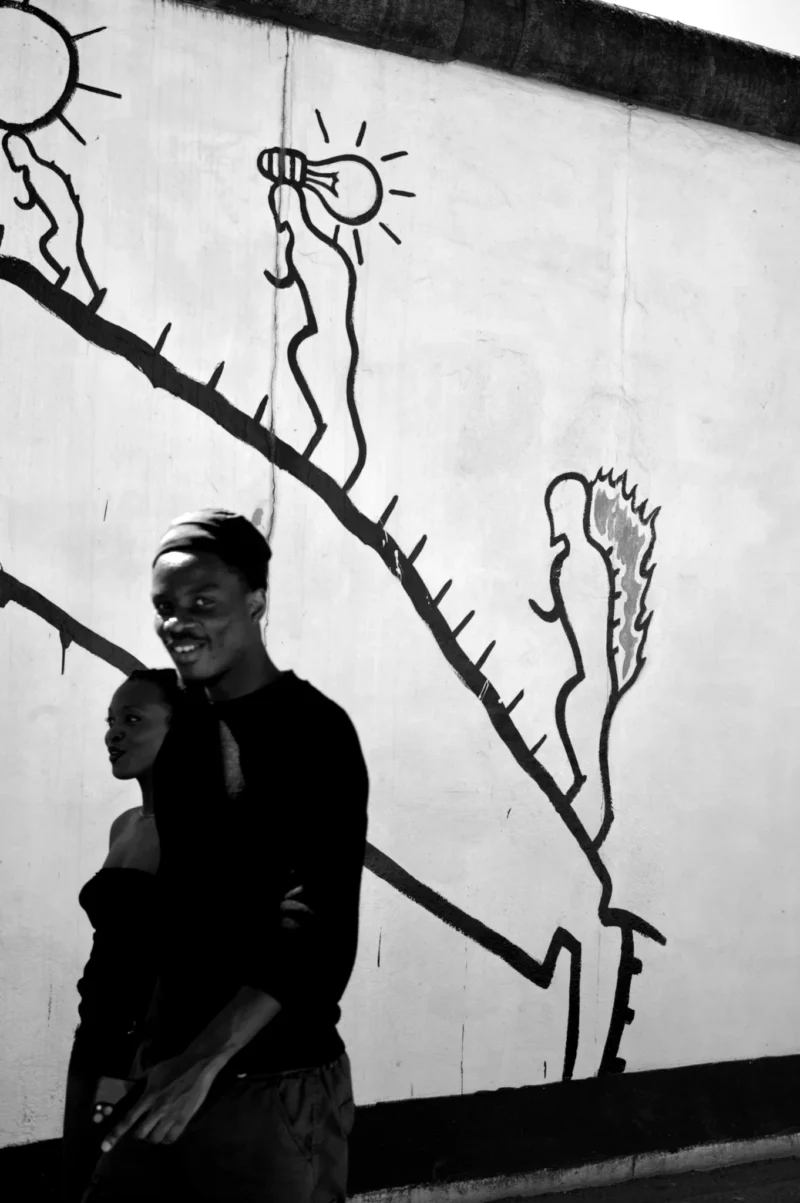 Thomas · 02/05/2026 · 1362 x 2048 Cette photographie en noir et blanc met en scène un homme masqué, sujet principal, marchant devant un mur tagué. La composition joue sur le contraste entre le personnage et les graffitis stylisés, évoquant des formes organiques et solaires. Le cadrage serré isole le sujet, créant une ambiance mystérieuse et urbaine.La lumière, naturelle et tamisée, accentue les ombres et les textures. Les couleurs, limitées au noir et blanc, renforcent le côté dramatique et artistique. Le style rappelle le street art et le cinéma noir, avec une ambiance à la fois sombre et poétique.