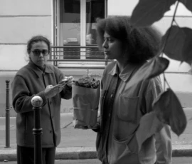 Thomas · 14/05/2026 · 3072 x 4096 Cette photographie en noir et blanc montre deux personnes en conversation sur un trottoir. Le sujet principal semble être l’échange entre eux, avec une composition centrée sur leurs visages et leurs gestes.Le cadrage est serré, mettant en valeur leurs expressions. La lumière est naturelle, douce et légèrement contrastée, créant des ombres subtiles. L’ambiance est calme et intime, renforcée par le style documentaire et réaliste.Les couleurs, bien que monochromes, jouent sur les nuances de gris, ajoutant une touche vintage et authentique à la scène.