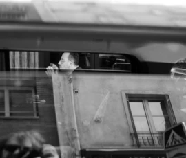 Thomas · 16/05/2026 · 3080 x 2048 Cette photographie en noir et blanc capture un homme penché hors d’un train en mouvement, s’accrochant à une porte. Le sujet principal est ce personnage, exprimant une urgence ou une précarité.La composition est dynamique, avec un cadrage serré sur le visage et le haut du corps. L’arrière-plan montre un bâtiment flou, renforçant le mouvement. La lumière est naturelle, contrastée, créant des ombres marquées. Les tons dominants sont les gris et noirs, typiques du noir et blanc, accentuant le côté dramatique.L’ambiance est tendue, presque désespérée, évoquant une scène de crise ou de danger. Le style est documentaire et réaliste.