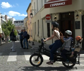 Bao · 4610 x 3418 Cette photographie montre une personne conduisant un vélo avec un enfant assis dans un siège arrière, sur une rue pavée. Le sujet principal est ce duo en mouvement.La composition met en valeur la scène urbaine avec des bâtiments en fond, dont un restaurant nommé "L'Espérance". Le cadrage est serré sur les cyclistes, mais inclut aussi des passants et une partie de l'architecture environnante.La lumière est naturelle et claire, évoquant une journée ensoleillée. Les couleurs dominantes sont les tons pastel des bâtiments, les teintes vives des vêtements et le bleu du ciel. L'ambiance est paisible et quotidienne, typique d'une ville européenne.