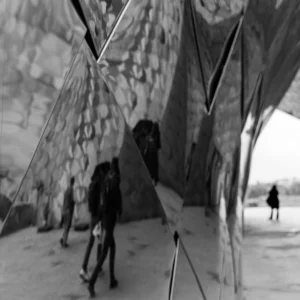 Laurence · 03/05/2026 · 900 x 900 Cette photographie en noir et blanc met en scène deux personnes marchant sous une structure architecturale géométrique et métallique. Le sujet principal est l'interaction entre les individus et l'espace reflétant, créant une ambiance mystérieuse et onirique. La composition joue sur les reflets et les angles, renforçant une impression de profondeur et de distorsion. Le cadrage serré et les jeux de lumière, bien que contrastés, accentuent les ombres et les reflets, ajoutant une touche dramatique. Le style est épuré et moderne, évoquant une ambiance contemplative et presque abstraite.