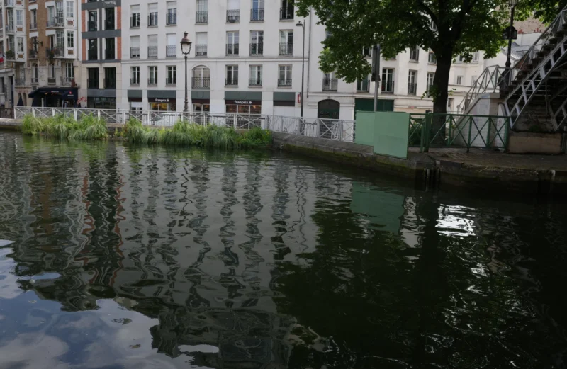 Bao · 5854 x 3809 Cette photographie capture un canal urbain calme. Le **sujet principal** est l’eau paisible reflétant les bâtiments et la végétation. La **composition** est épurée, avec une ligne horizontale marquée par le niveau de l’eau et une perspective légèrement inclinée.Le **cadrage** est serré, centré sur le canal et ses bords. La **lumière** semble naturelle, douce et tamisée, créant des ombres discrètes. Les **couleurs dominantes** sont des tons verts (végétation), gris (bâtiments, eau) et quelques touches de blanc.Le style est **réaliste et minimaliste**, évoquant une **ambiance sereine et presque mélancolique**, typique d’un espace urbain apaisant.