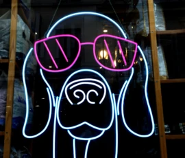 Bao · 6024 x 4016 Cette photographie met en scène un chien stylisé en néon, sujet principal. La composition est épurée, centrée sur l’animal aux yeux exagérés et aux lunettes roses. Le cadrage est serré, isolant le motif lumineux.Les couleurs dominantes sont le bleu électrique du chien et le rose vif des lunettes, créant un contraste saisissant. La lumière, artificielle et néon, donne un aspect futuriste et vibrant.Le style est minimaliste et graphique, évoquant une ambiance nocturne et moderne, presque rétro-futuriste. L’éclairage accentue une atmosphère dynamique et légèrement onirique.